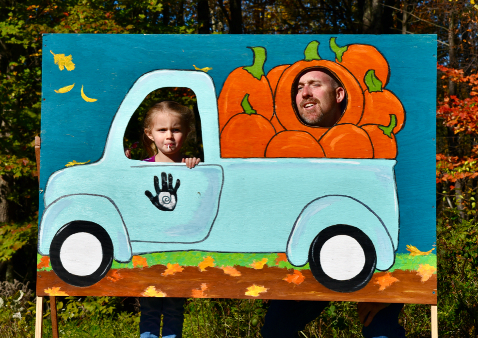 A parent and their child standing behind a photo stand-in of a truck full of pumpkins